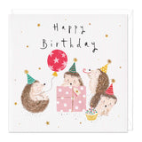 Hedgehog Party Happy Birthday Card features 4 hedgehogs with party hats on having the time of their lives! One is holding a red balloon, one popping his head out of a present, another tooting a party blower the other is eyeing up a delicious looking cupcake. There are gold glitter stars surrounding the scene and  gold foiled pom poms on the party hats.